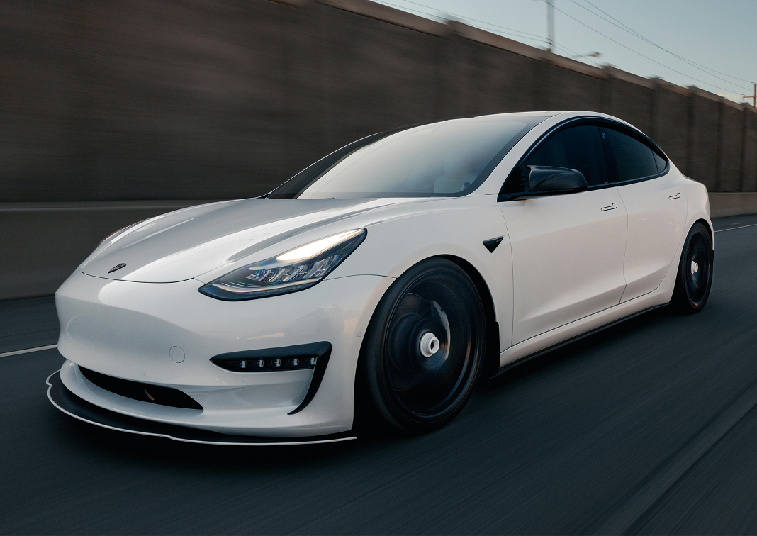 Білий седан Tesla Model 3 з чорними колесами їде по шосе біля бетонної стіни.